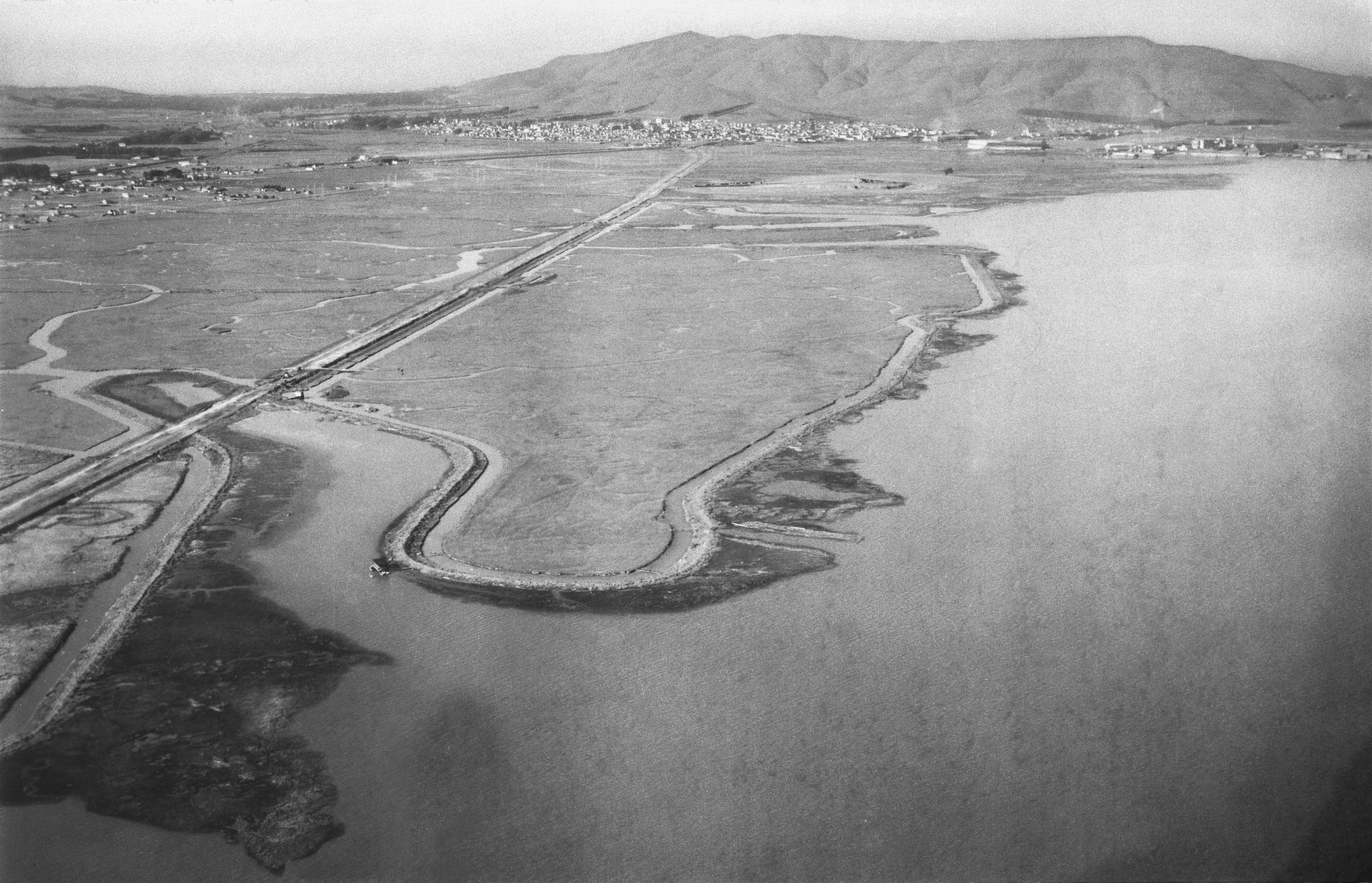 From attached handwritten note: “View N. Site of Mills Field 03542 January 1927”. Black and white copy negative depicting oblique aerial view over Mills Field Municipal Airport of San Francisco, facing north with San Francisco Bay at right and old Bayshore Highway at left; structures visible in background against San Bruno Mountain; Mills Field Municipal Airport of San Francisco; photo taken January 1927; inscribed on white border, right: “JAN 1927”.