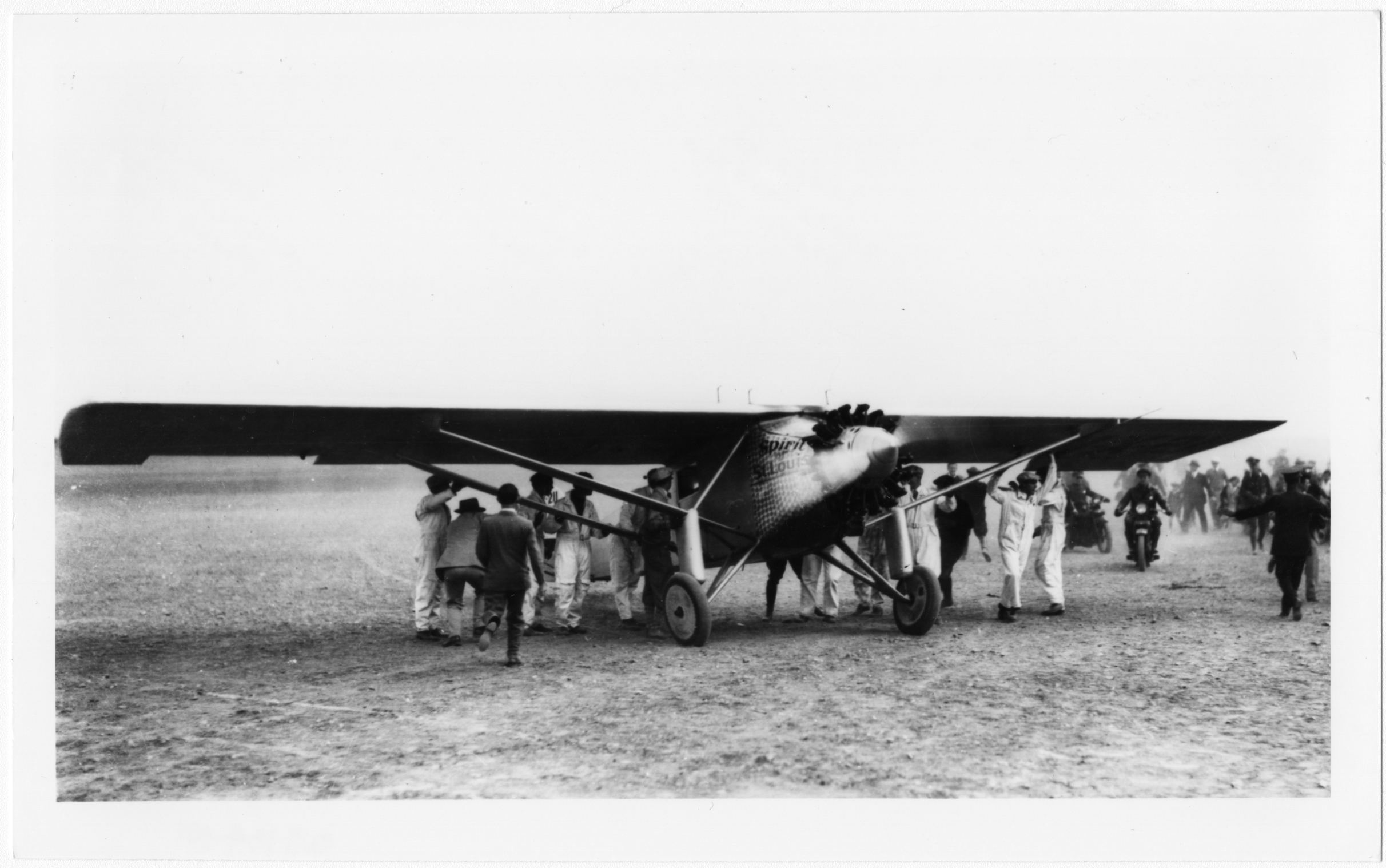 Black and white photographic copy print of Charles Lindbergh arriving in the Spirit of St. Louis on Mills Field Municipal Airport, San Francisco, September 16, 1927. Image depicts airplane from front at oblique angle with crews in white coveralls under airplane wings. propeller is in motion, with men in suits approaching airplane. Police officers on motorcycles and crowd of people in background.